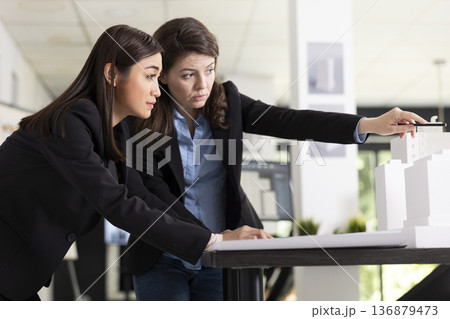 Focused diverse women analyze blueprints and scale model, preparing for upcoming construction site visit. Female architects making necessary adjustments to ensure building project meets requirements. 136879473