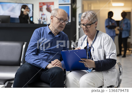 Caucasian general practitioner sits with retired man, explaining blood work results in reassuring manner. Female doctor and male pensioner patient having medical conversation in clinic lobby. 136879478