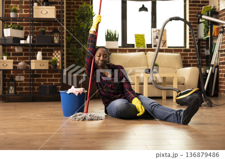 Exhausted woman sits on floor with mop after cleaning, showing fatigue from household chores. Black housewife wearing yellow gloves, leans on cleaning stick, resting from domestic duties at home. Exhausted woman sits on floor with mop after cleaning, showing fatigue from household chores. Black housewife wearing yellow gloves, leans on cleaning stick, resting from domestic duties at home. 136879486
