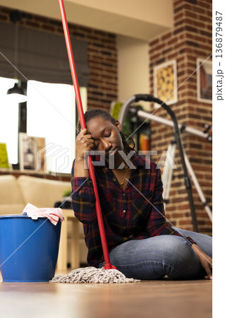 African american woman sits on floor, leaning on mop handle with eyes closed, looking tired after daily cleaning chores. Weary housewife takes break from mopping in brick wall living room. African american woman sits on floor, leaning on mop handle with eyes closed, looking tired after daily cleaning chores. Weary housewife takes break from mopping in brick wall living room. 136879487