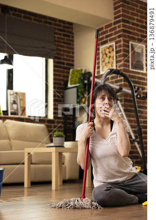 Caucasian woman sits on floor with mopping stick, yawning and covering her mouth, exhausted after long day of housecleaning. Young lady takes moment to rest before completing household chores. Caucasian woman sits on floor with mopping stick, yawning and covering her mouth, exhausted after long day of housecleaning. Young lady takes moment to rest before completing household chores. 136879491