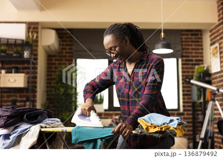 Smiling black housewife using electric iron to press clean shirt, removing wrinkles after laundry. Cheerful and focused, african american woman completes household chores in modern apartment. 136879492