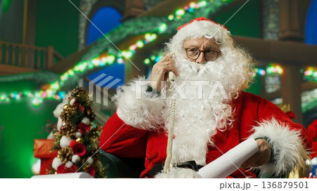 Santa Claus answering phone call, responding to kids requesting gifts. Person impersonating Santa Claus talking with children about Christmas gifts using landline telephone, camera A 136879501