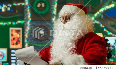 Happy man dressed as Santa Claus making list of kids who has been naughty or nice before sending gifts. Joyous person preparing to travel all over the world, deliver toys to children, camera A Happy man dressed as Santa Claus making list of kids who has been naughty or nice before sending gifts. Joyous person preparing to travel all over the world, deliver toys to children, camera A 136879511