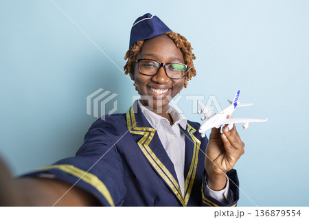 Black female air hostess playfully poses for selfie while holding model aircraft. Cheerful cabin crew member enjoys documenting experience in aviation industry in blue background studio. Black female air hostess playfully poses for selfie while holding model aircraft. Cheerful cabin crew member enjoys documenting experience in aviation industry in blue background studio. 136879554