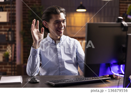 Happy businessman waves hand at screen, joining computer video conference from home. Male professional participates in online meeting, negotiation and remote teamwork using digital technology. Happy businessman waves hand at screen, joining computer video conference from home. Male professional participates in online meeting, negotiation and remote teamwork using digital technology. 136879556