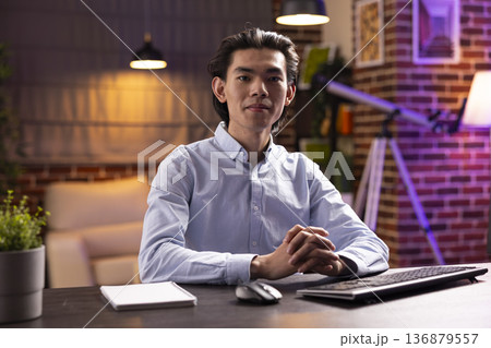 Confident professional man and entrepreneur sits at desk with computer keyboard, looking at camera. Portrait of focused asian male executive and leader working remotely in modern apartment. 136879557