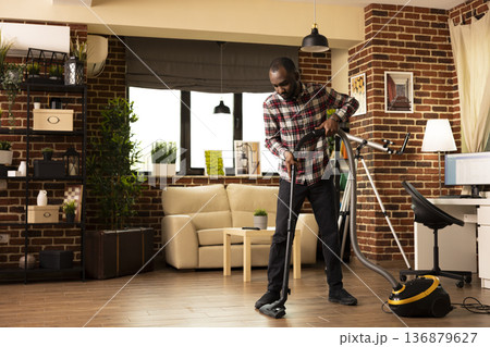 African American male homeowner vacuuming living room floor in bright apartment. Young black man holding household appliance, focused on keeping brick wall home clean and tidy during weekend chores. African American male homeowner vacuuming living room floor in bright apartment. Young black man holding household appliance, focused on keeping brick wall home clean and tidy during weekend chores. 136879627