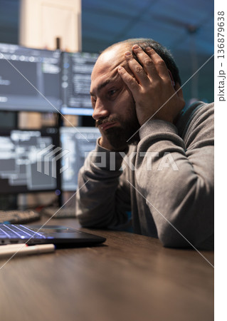 Overworked programmer rests head in palms during difficult coding session. Male developer analyzes complex data on laptop with exhausted expression, represents programming stress and technical 136879638