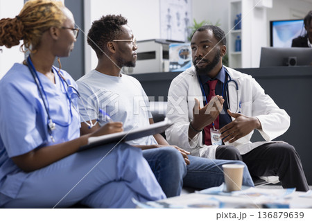 Black male doctor in lab coat speaks with patient during clinic appointment, reviewing documents and discussing health concerns. Female nurse takes notes as physician explains treatment information. Black male doctor in lab coat speaks with patient during clinic appointment, reviewing documents and discussing health concerns. Female nurse takes notes as physician explains treatment information. 136879639