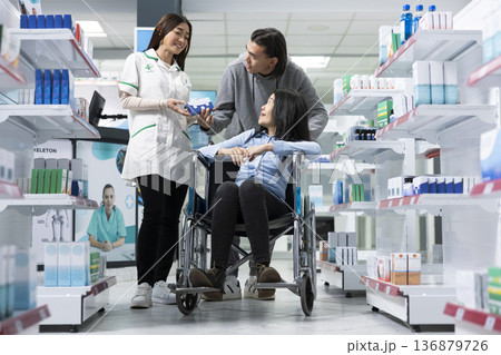 Female pharmacist provides inclusive healthcare service to woman in wheelchair and her companion. Focus on pharmacy accessibility, patient support and professional medical guidance in store. Female pharmacist provides inclusive healthcare service to woman in wheelchair and her companion. Focus on pharmacy accessibility, patient support and professional medical guidance in store. 136879726