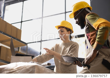 Two employees in coveralls and hardhat manage inventory control on laptop, product handling and packing goods for dispatch. Online shopping shipment in small scale logistics depot. 136879757