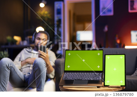 Black modern guy enjoys leisure time next to copy space at his home, using gadgets to watch a podcast or a movie with headset. Young casual adult relaxing remotely in the living room. Black modern guy enjoys leisure time next to copy space at his home, using gadgets to watch a podcast or a movie with headset. Young casual adult relaxing remotely in the living room. 136879780