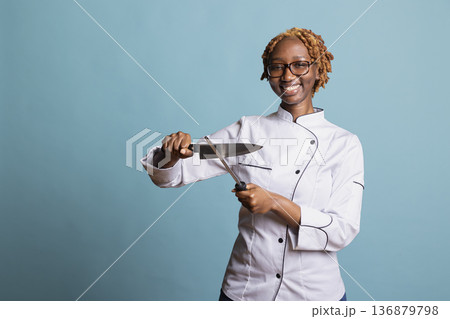 Smiling female chef using honing steel to sharpen knife before preparing food. Professional cook stands confidently against isolated background, enjoying art and precision of culinary routine. 136879798