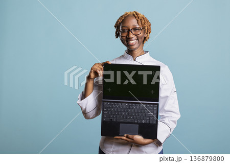 Joyful female chef in uniform presents laptop with green screen over isolated blue background. Professional woman cook displays chromakey template for restaurant promotions and culinary advertising. Joyful female chef in uniform presents laptop with green screen over isolated blue background. Professional woman cook displays chromakey template for restaurant promotions and culinary advertising. 136879800