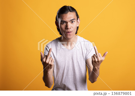 Asian man looks directly at camera with anger and frustration, raising hands in bold, rebellious stance. Furious male individual expressing defiance, tension and emotional strength in studio. 136879801