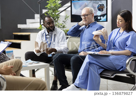 Caucasian man points to neck and shoulder pain during consultation with medical specialists, helping guide diagnosis and treatment for upper body discomfort. Diverse physicians speaking with patient. Caucasian man points to neck and shoulder pain during consultation with medical specialists, helping guide diagnosis and treatment for upper body discomfort. Diverse physicians speaking with patient. 136879827