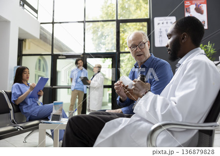 African american doctor explains prescription details and proper pill usage to senior man in hospital waiting room. Medical consultation emphasizes treatment understanding and recovery support. African american doctor explains prescription details and proper pill usage to senior man in hospital waiting room. Medical consultation emphasizes treatment understanding and recovery support. 136879828