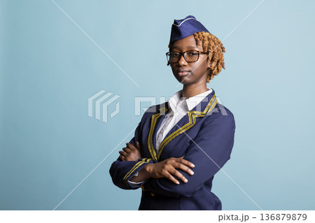 Portrait of black air hostess in blue and gold uniform crossing arms confidently. Young woman in glasses stands against isolated studio background, representing professional airline services. Portrait of black air hostess in blue and gold uniform crossing arms confidently. Young woman in glasses stands against isolated studio background, representing professional airline services. 136879879