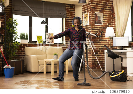 Positive housewife dances in living room, cleaning floor with vacuum and maintaining cleanliness in brick wall apartment. Happy young woman wears headphones, listening to music and vacuuming at home. Positive housewife dances in living room, cleaning floor with vacuum and maintaining cleanliness in brick wall apartment. Happy young woman wears headphones, listening to music and vacuuming at home. 136879930