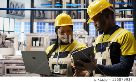 Solar panels factory engineering team analyzes performance data on laptop to predict gear failures. Teamworking photovoltaics plant colleagues following maintenance procedures, camera A Solar panels factory engineering team analyzes performance data on laptop to predict gear failures. Teamworking photovoltaics plant colleagues following maintenance procedures, camera A 136879937