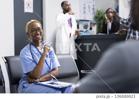 African American female nurse sits in clinic reception area, getting ready for duties and patient care. Cheerful medical professional looks at camera with confidence while waiting in hospital lobby. 136880021