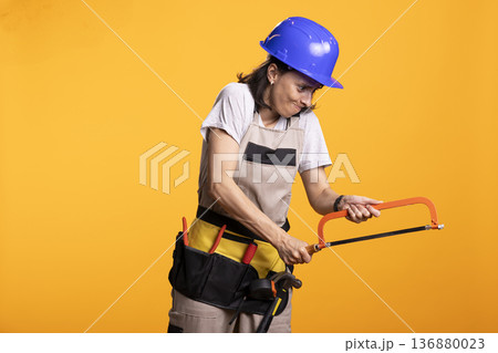 Female builder in hardhat and tool belt grips handsaw with focused expression while simulating cutting action. Renovation worker in studio demonstrates DIY effort against bold yellow background. 136880023