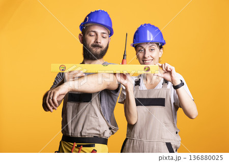 Male and female builders smile while holding measuring level together, demonstrating teamwork and accuracy at renovation site. Construction workers collaborating on building plans with tools in hand. 136880025