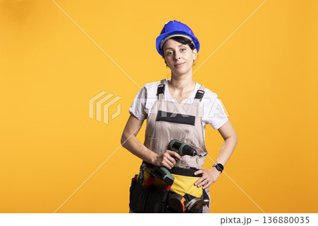 Female adult stands against isolated background wearing construction workwear and safety helmet, holding drill gun with confidence. Female renovator demonstrates home improvement professionalism Female adult stands against isolated background wearing construction workwear and safety helmet, holding drill gun with confidence. Female renovator demonstrates home improvement professionalism 136880035