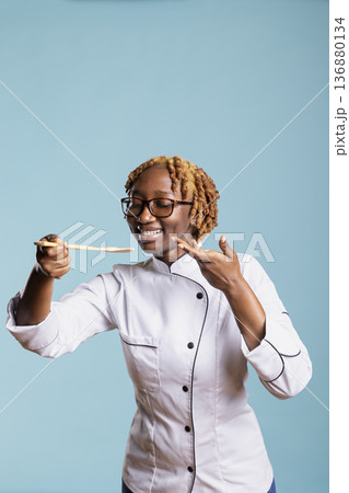 Joyful female chef in uniform tasting food using wooden spoon against blue background. Professional cook expressing satisfaction and creativity, showing passion and excellence for culinary arts. 136880134