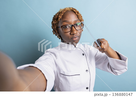 African American professional cook playfully pointing kitchen knife toward camera, recording culinary video. Confident female chef shows seriousness in preparation for gourmet challenge. 136880147