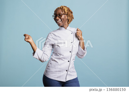 Excited professional cook in uniform celebrates culinary accomplishments, expressing happiness and pride in preparing gourmet dishes. Young female chef dancing and having fun in studio. Excited professional cook in uniform celebrates culinary accomplishments, expressing happiness and pride in preparing gourmet dishes. Young female chef dancing and having fun in studio. 136880150