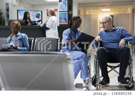 African american nurse seated with elderly male wheelchair user, confirming medical information before doctors appointment. Black female medic with clipboard, explaining recovery plan to senior man. 136880156