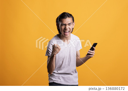 Joyful asian man holds smartphone and celebrates with clenched fist after receiving good news. Handsome model expressing triumph over winning lottery or receiving successful business deal notification 136880172