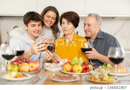 Senior woman and man celebrating family holiday with their son and his young wife at kitchen table, looking at photos on smartphone Senior woman and man celebrating family holiday with their son and his young wife at kitchen table, looking at photos on smartphone 136881867
