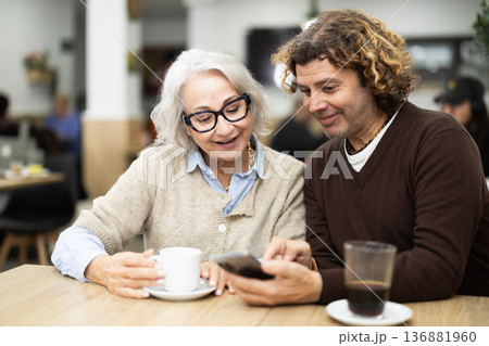 Happy elderly lady meeting with middle-aged man talking and dining in restaurant, looking at photos on mobile phone Happy elderly lady meeting with middle-aged man talking and dining in restaurant, looking at photos on mobile phone 136881960