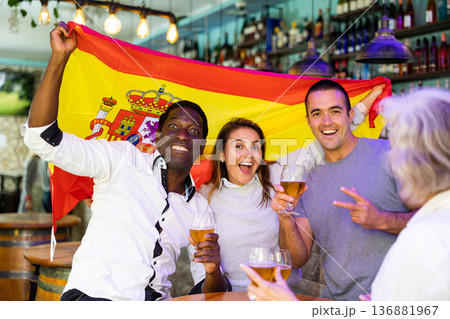 Happy multiracial sport fans holding and wrapping in a flag of Spain and supporting national football team in the bar 136881967