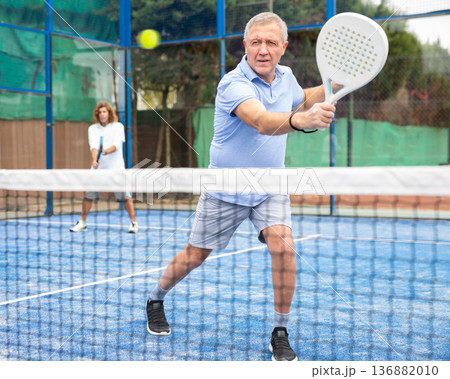 Serious old man tennis player playing padel during friendly match on outdoor court in spring Serious old man tennis player playing padel during friendly match on outdoor court in spring 136882010