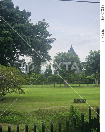 A majestic view of the ancient Prambanan Hindu temple towering behind lush green trees and a wide grassy field under a cloudy sky, showcasing Indonesia's historic stone architecture and heritage site. 136882035