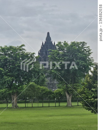 A majestic view of the ancient Prambanan Hindu temple towering behind lush green trees and a wide grassy field under a cloudy sky, showcasing Indonesia's historic stone architecture and heritage site. 136882038