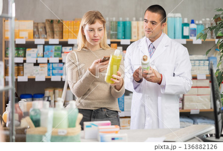 Young woman in pharmacy scans QR code on hair loss shampoo, man pharmacist in background 136882126