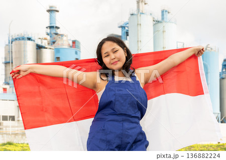 Positive girl in overalls with the Indonesia flag in her hands against the background of chimneys of modern factory 136882224