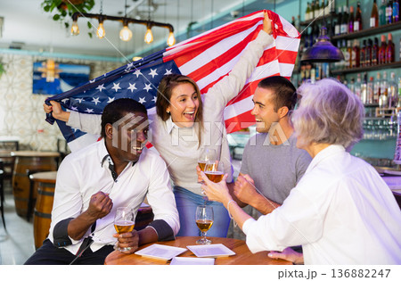 Cheerful multiracial footbal fans waving the flag of USA while drinking beer and watching tournament in sport bar 136882247