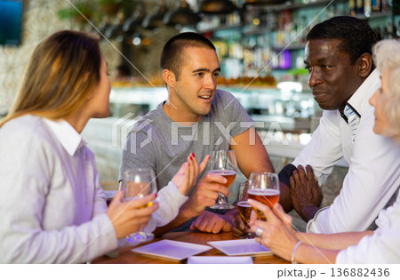 Portrait of smiling females and males drinking beer and having fun conversation in bar. Focus on young man 136882436