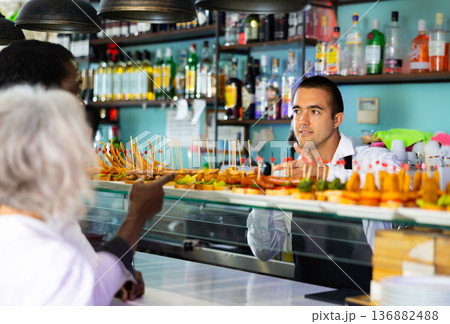 Bartender standing at counter in bar, selling fresh sandwiches pinchos to client 136882488