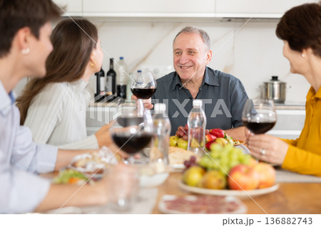 Senior man at kitchen table with wife, grown son and daughter, enjoying conversation at festive table with wine and snacks 136882743