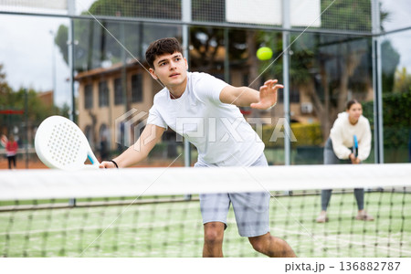 Sportive young man holding padel racquet in hand and ready to return ball while playing in court Sportive young man holding padel racquet in hand and ready to return ball while playing in court 136882787
