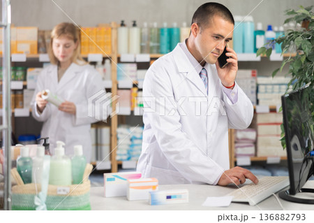 Man pharmacist works at computer and talks on phone. Girl assistant in background 136882793