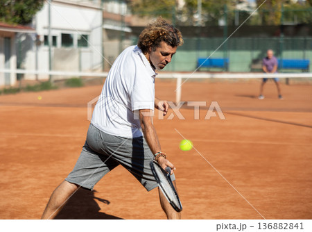 Back view of active male player hitting ball with racket on tennis court on blurred background of opponents 136882841