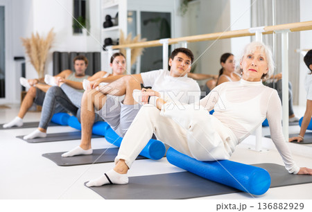 Elderly woman practicing pilates with roller on gray mat in gym room 136882929